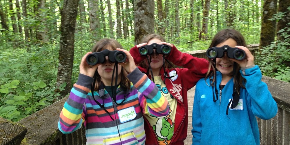 Three students hold binoculars to their eyes on the Refuge boardwalk.